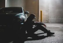 5 sygnałów, że jesteś za bardzo zmęczony na prowadzenie, choć wydaje ci się inaczej Zmęczony kierowca siedzi na asfalcie oparty o zaparkowane auto o zmierzchu