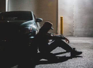 5 sygnałów, że jesteś za bardzo zmęczony na prowadzenie, choć wydaje ci się inaczej Zmęczony kierowca siedzi na asfalcie oparty o zaparkowane auto o zmierzchu